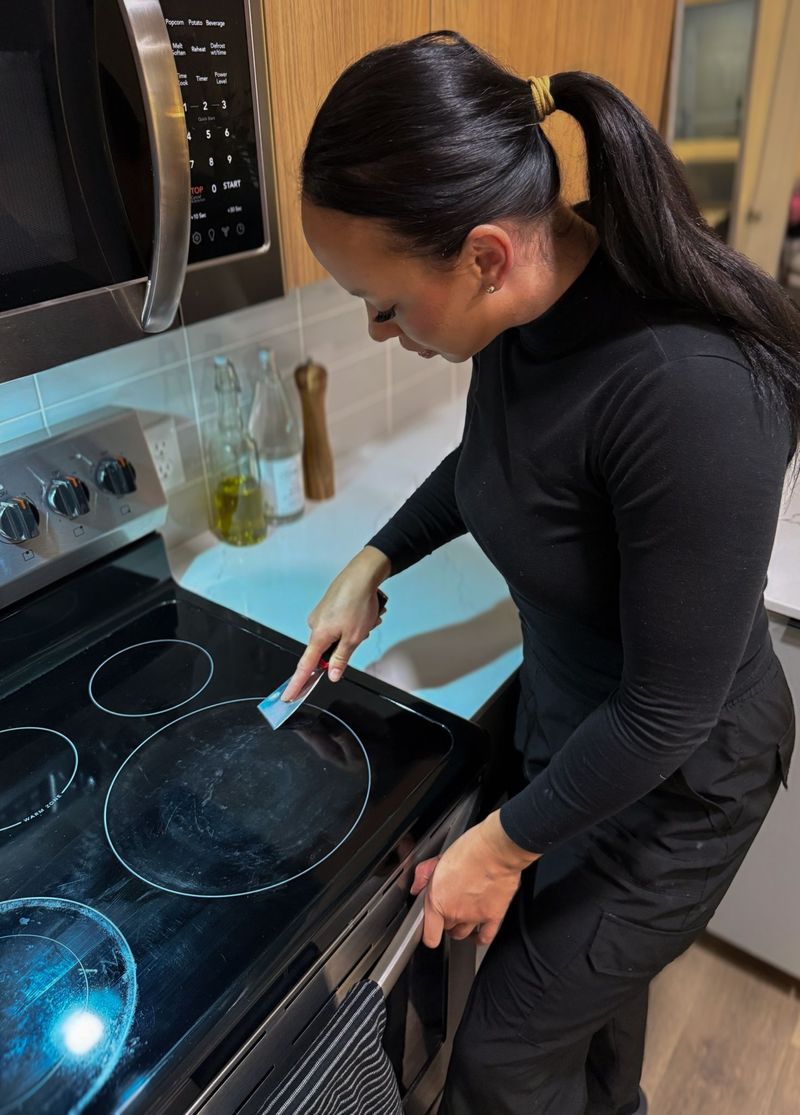 A professional cleaner stands in a kitchen, scraping residue from a glass stovetop using a cleaning tool, highlighting detailed appliance maintenance and deep cleaning in a modern residential environment.
