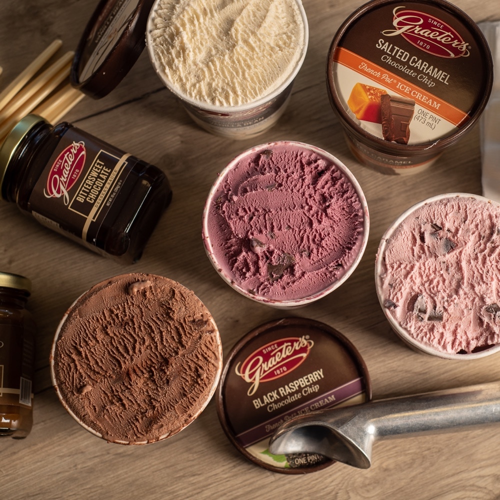 Four different pints of Graeter's Ice cream open with a metal ice cream scoop laid in front, Graeter's Butterscotch Dessert Sauce & Bittersweet Dessert Sauce lay on their sides and a stack of disposable ice cream bowls that say 'Graeter's since 1970' lay on their side.