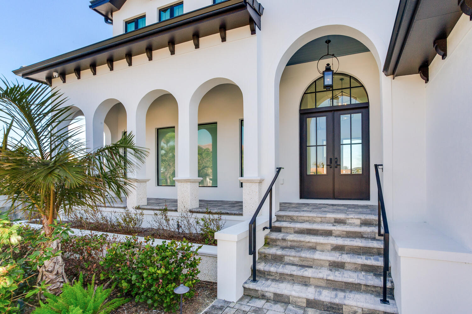 Mediterranean-style AVENTIS custom home entry featuring arched windows, a decorative front door, tile roof accents, and elegant coastal landscaping.