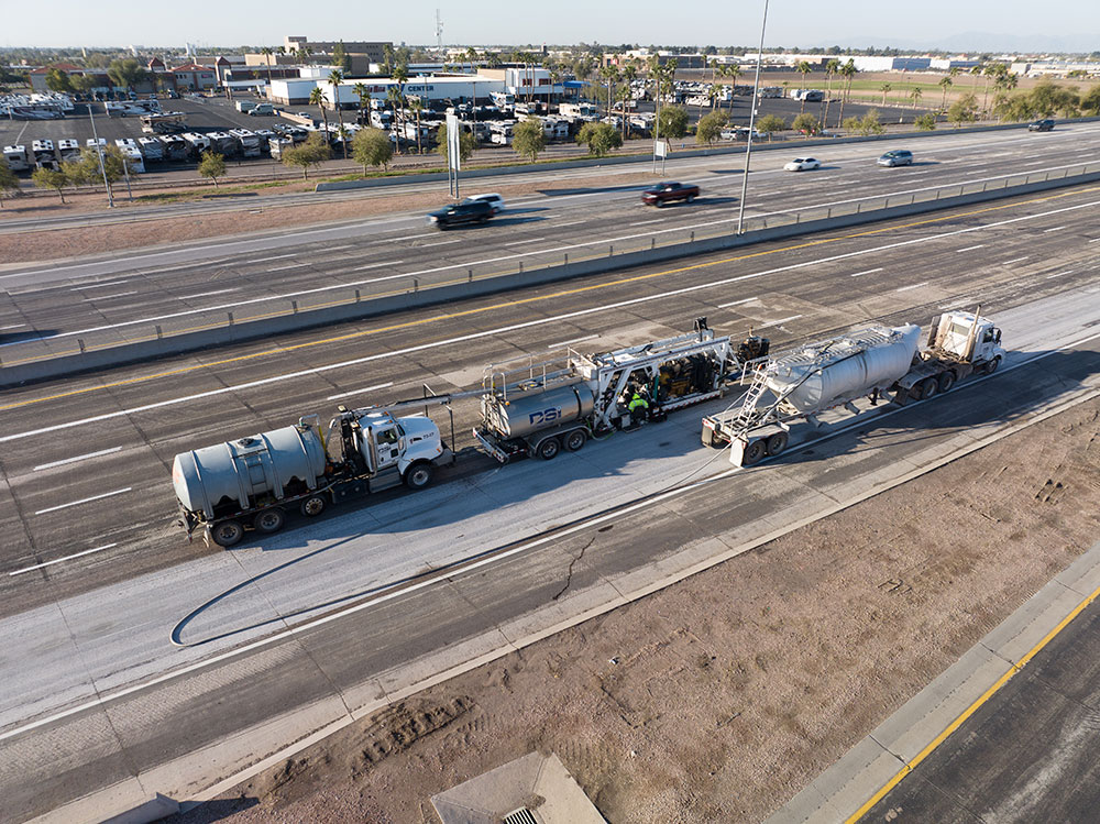 Elevated side-angle aerial photo capturing an entire DSI diamond grinding assembly staged along a concrete highway lane. The multi-unit setup features a front water tanker, the central diamond grinding rig with operators visible on the side platform, and a rear slurry vacuum/tanker truck, with an adjacent freeway running parallel in the background.