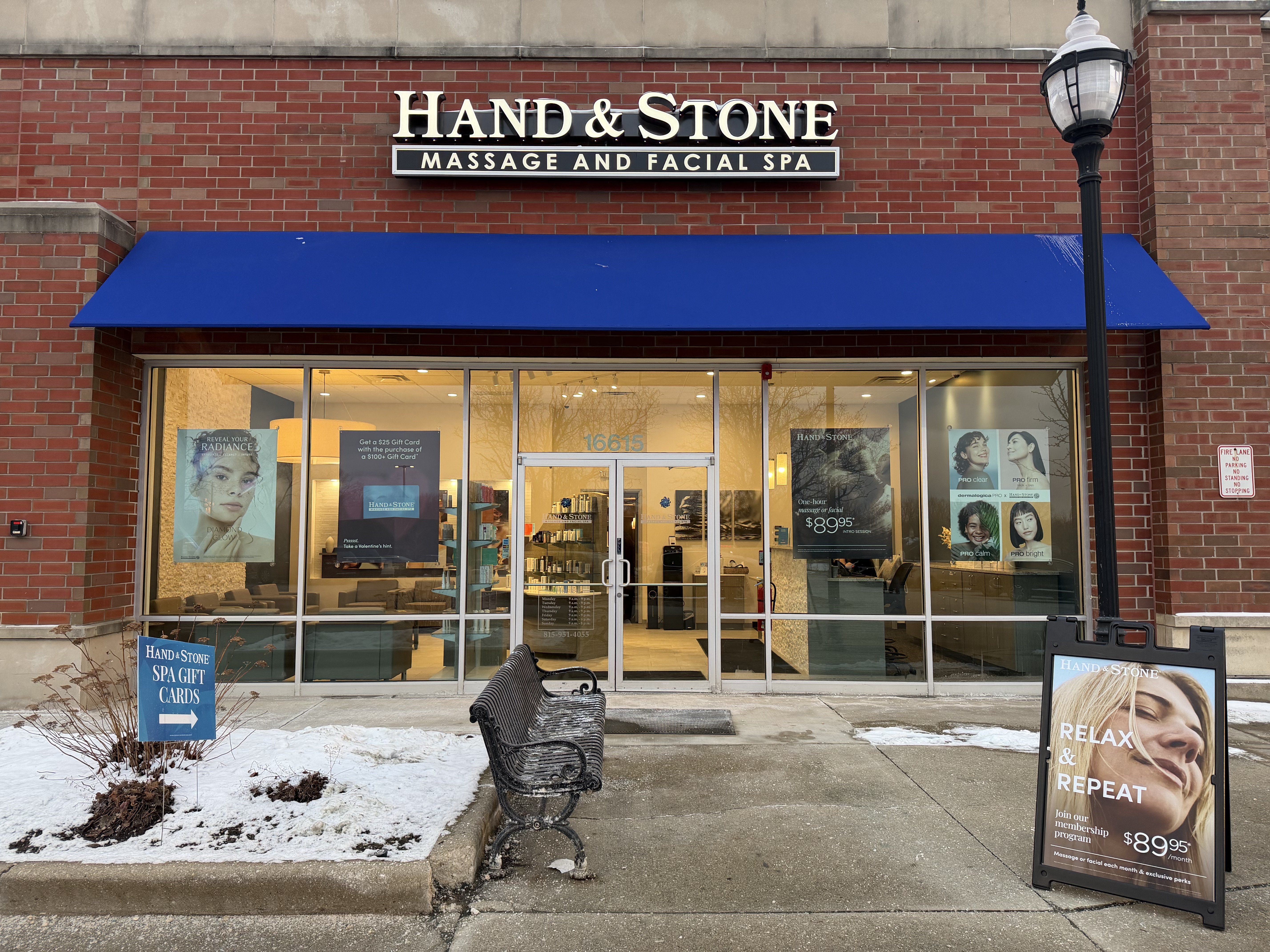 Brick storefront with prominent Hand & Stone signage in white serif lettering. Blue awning extends over aluminum-framed glass entrance. Large promotional windows display service posters. Black metal bench and A-frame membership sign on concrete sidewalk. Snow-dusted landscaping with blue directional sign for gift cards.