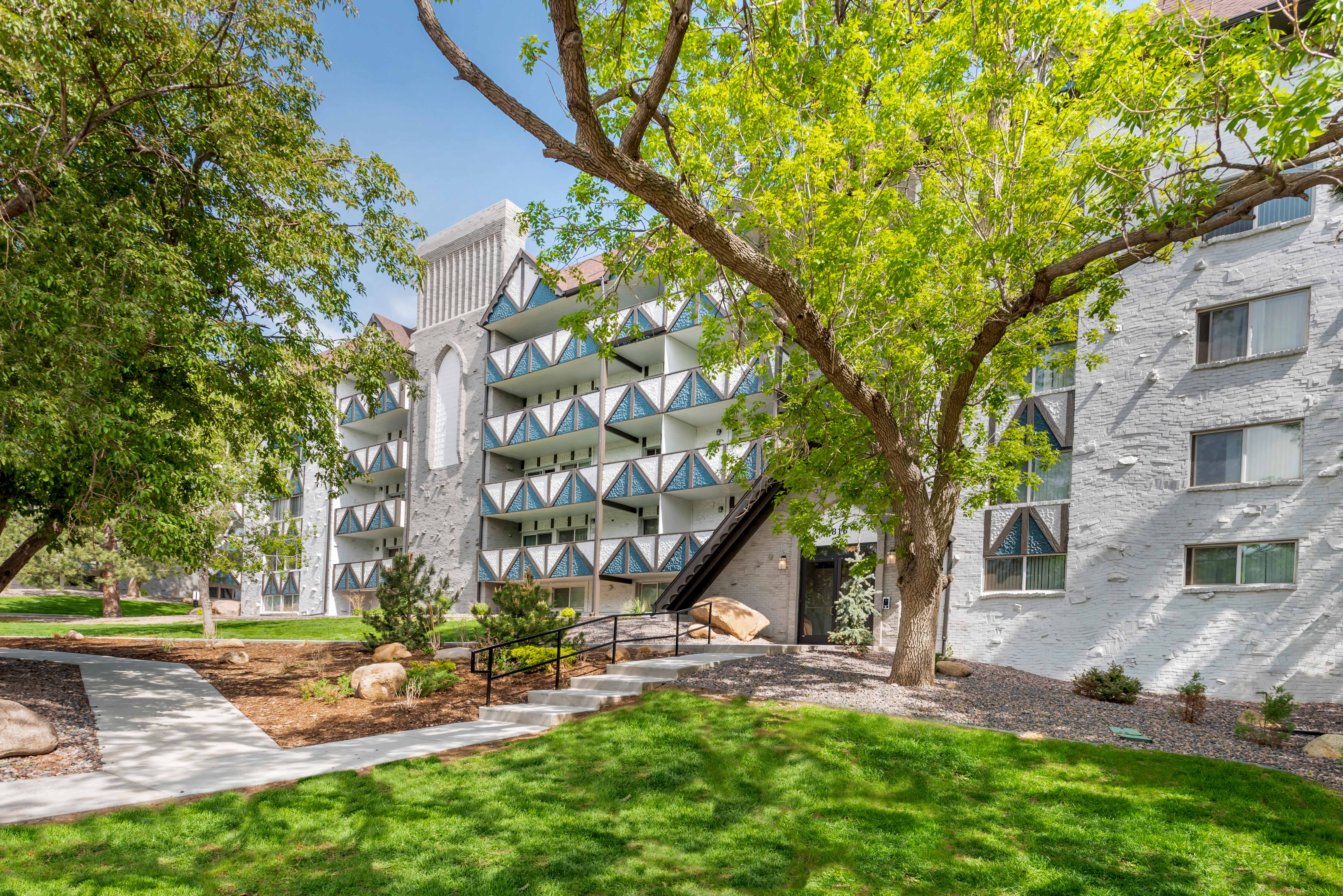 White, blue, and brown building exterior with geometric patterned balconies, trees, and grass.