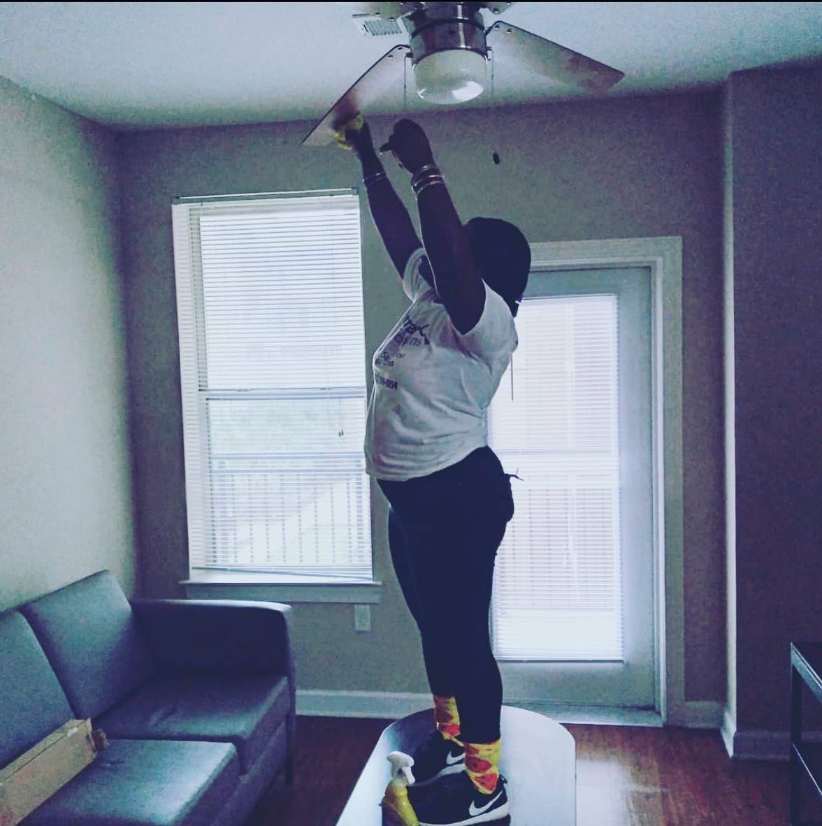 A woman is standing on a table and cleaning the blades of a ceiling fan with a yellow cloth.