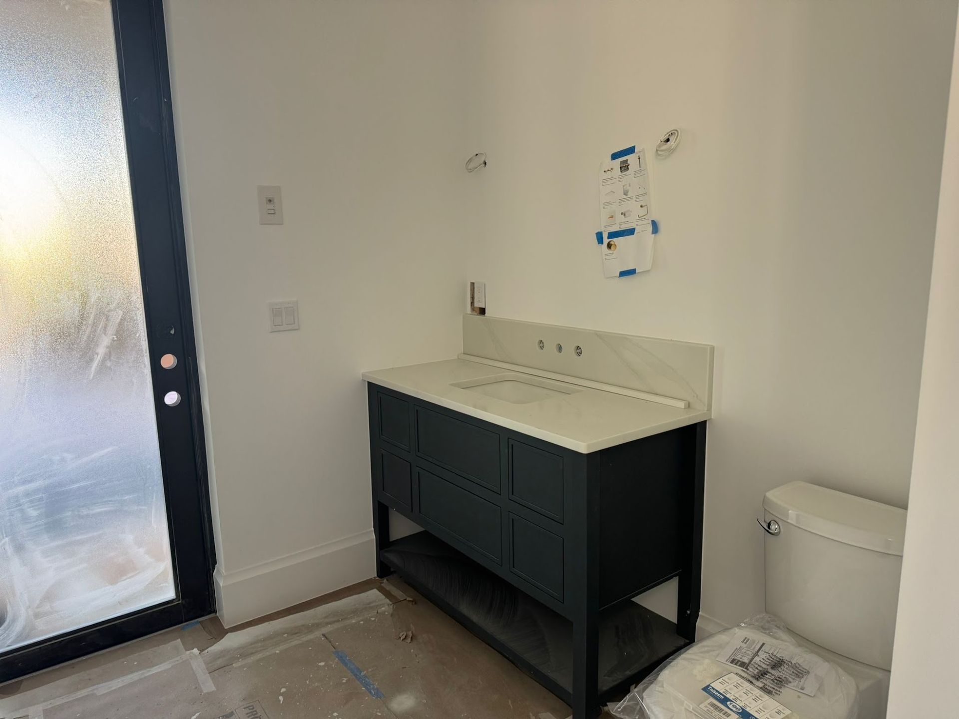 Partially completed bathroom installation with a dark vanity cabinet, white countertop sink, and nearby toilet. Walls are unfinished with fixture openings, and protective coverings remain on the floor during renovation.