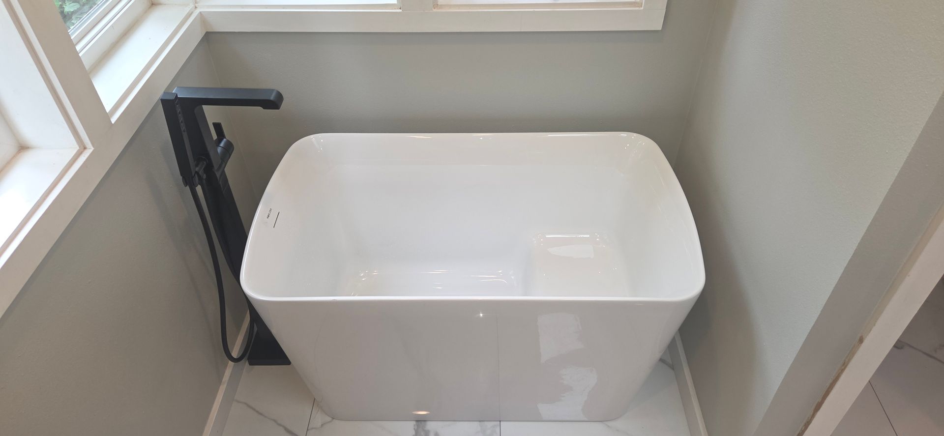 Freestanding white bathtub installed in a corner bathroom space beside windows, paired with a black  
