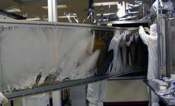 Technicians in full cleanroom suits operating DSI's proprietary MicroDyn® magnetron sputtering deposition system. The octagonally-shaped chamber with rotating drum enables uniform coating of complex geometries with superior density and durability compared to traditional evaporation methods. System produces coatings up to 30 square feet per batch run with extreme thermal resistance up to 1100°C.