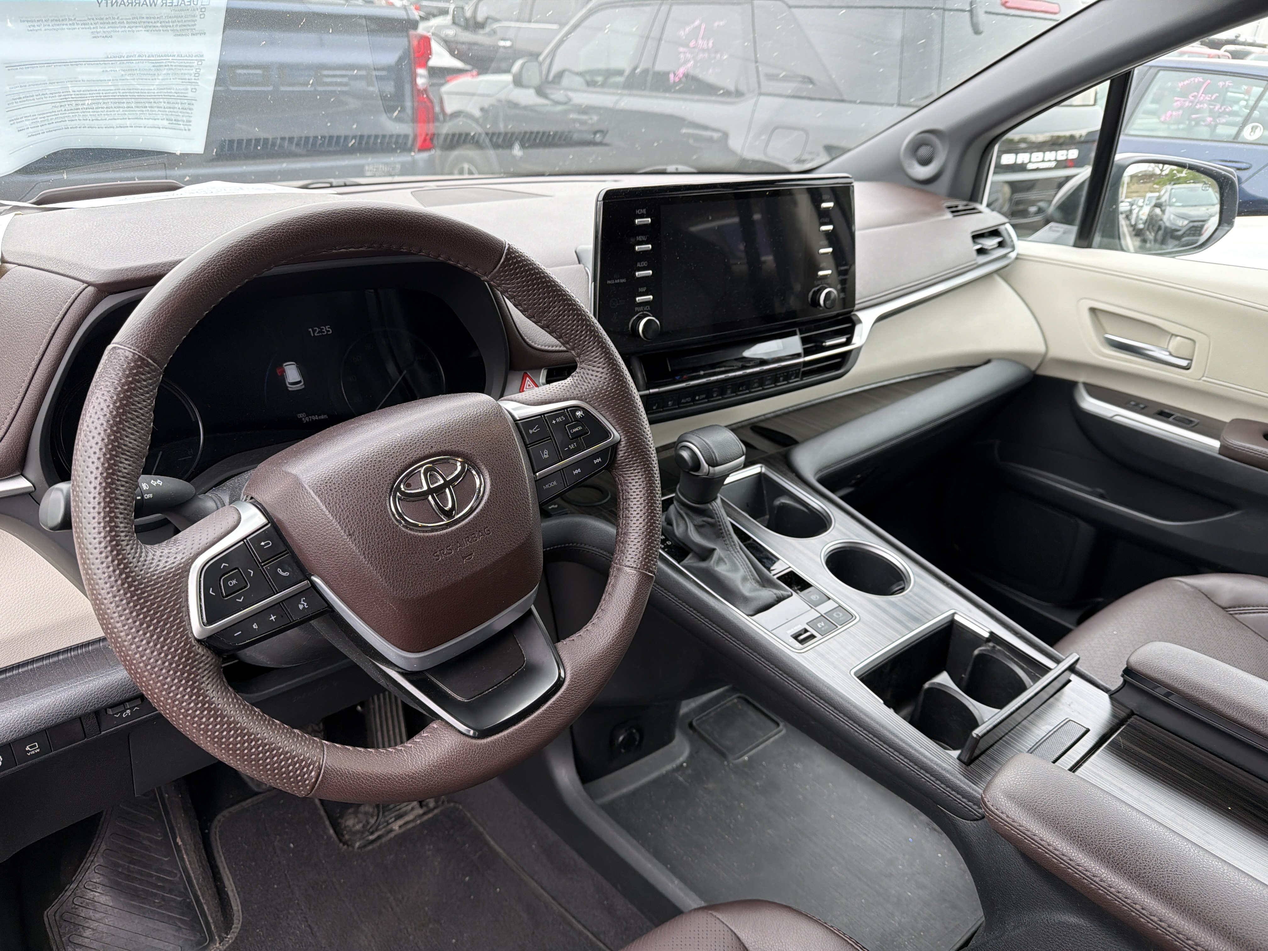 The interior of a modern car, showcasing the dashboard, steering wheel, center console, and seats. The dashboard features a large touchscreen infotainment system and a digital instrument cluster. The steering wheel is leather-wrapped with integrated controls. The center console includes a gear shifter, cup holders, and storage compartments. The seats are upholstered in brown leather. The overall aesthetic is luxurious and technologically advanced.