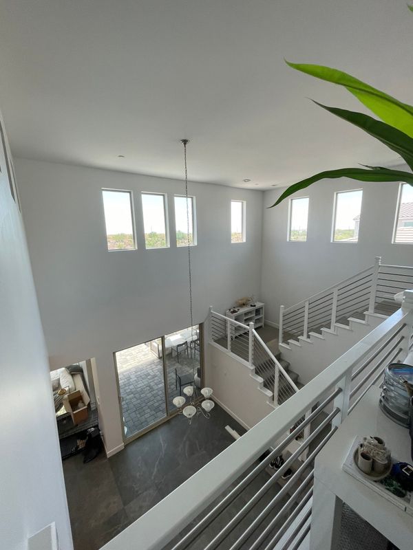 A modern open interior with high ceilings, multiple windows, and a staircase with white railings, overlooking a lower living area with chandelier lighting and clean, minimalist design.