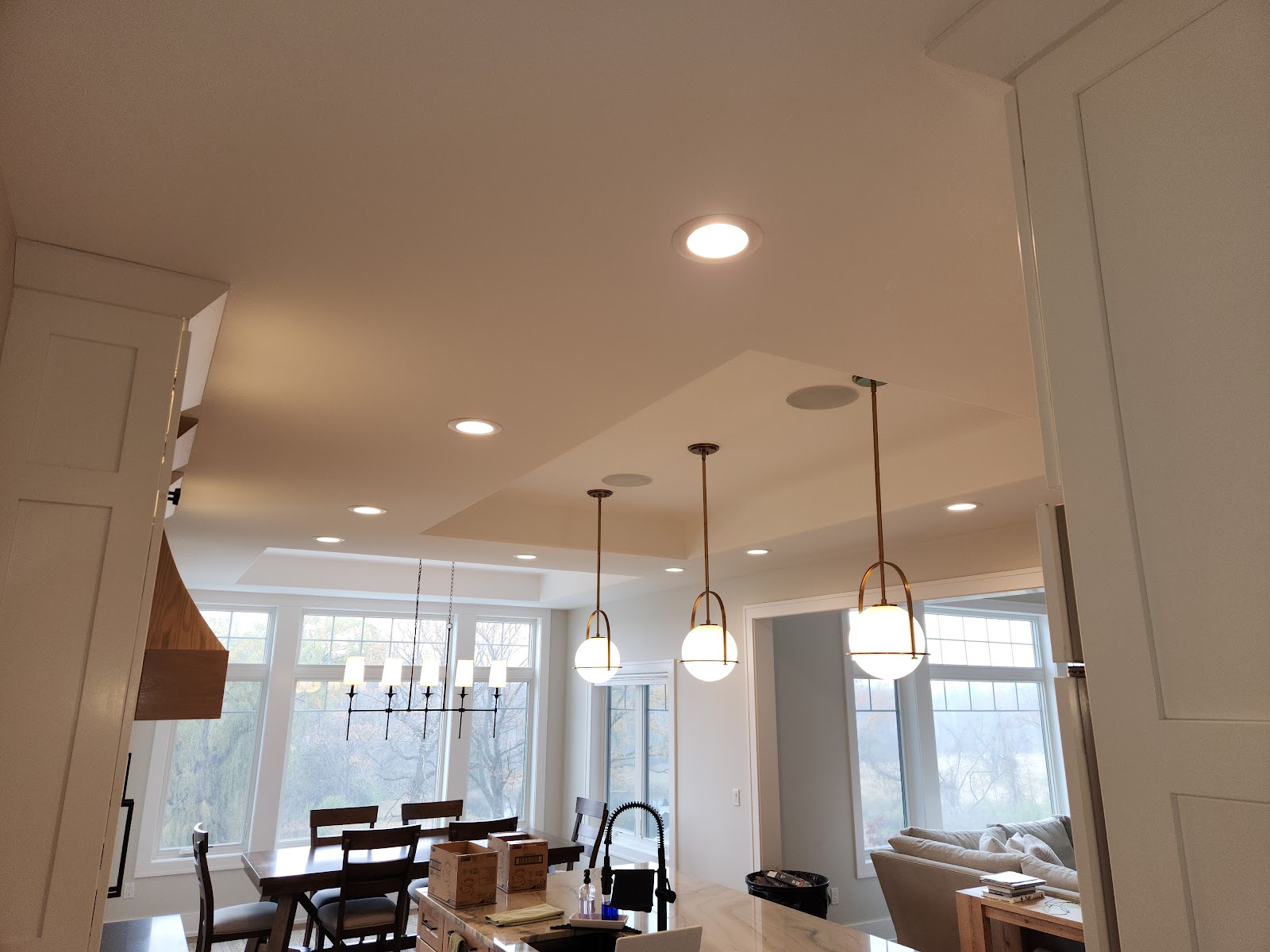 A modern kitchen and dining area with large windows overlooking a wooded landscape. The kitchen island features a marble countertop with a sink and faucet. Several pendant lights hang from the ceiling, along with a chandelier over the dining table. The dining area has a large wooden table with chairs. In the background, a living room area is visible with a sofa and side table.
