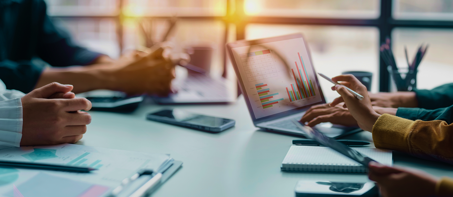 Close-up of a meeting room table, with hands pointing at a graph on a laptop.