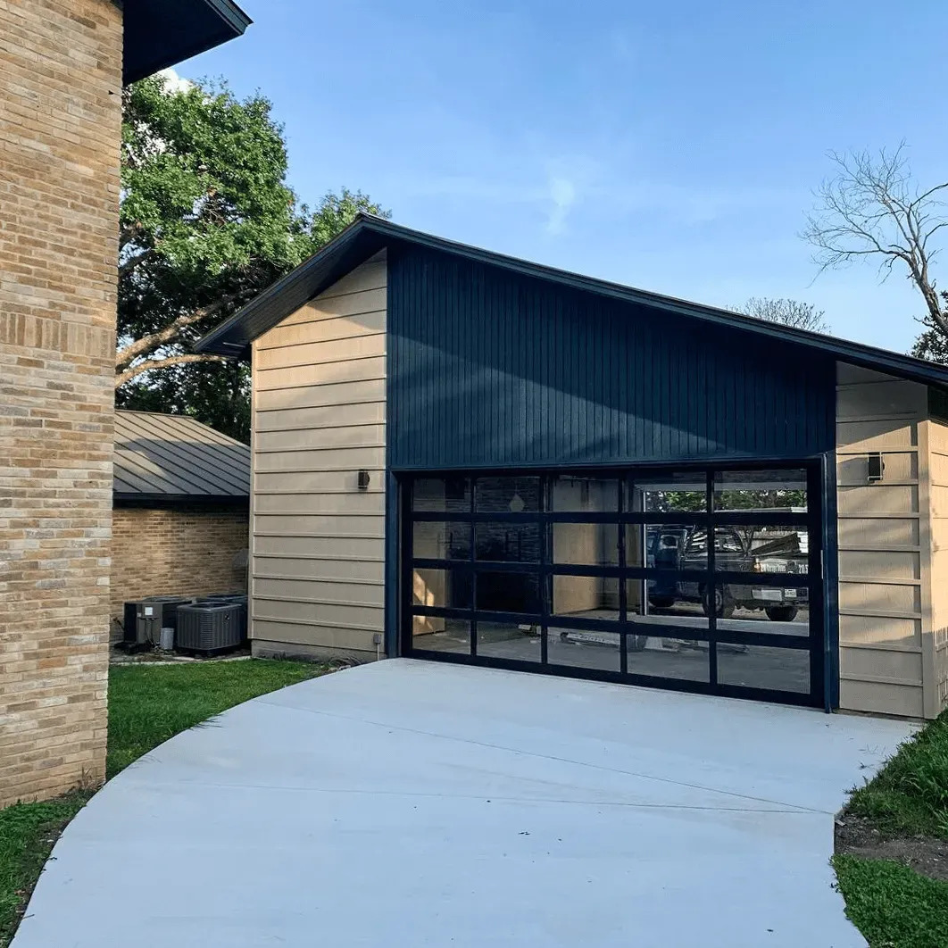 A detached modern garage features a black-framed glass garage door with multiple transparent panels, beige siding, dark trim, and a curved concrete driveway surrounded by green landscaping and lawn.