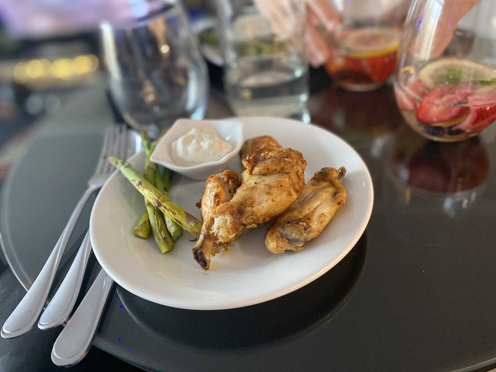 A close-up shot of a white plate filled with seasoned chicken wings and asparagus. The chicken wings appear golden brown and crispy, with visible seasoning. The asparagus spears are grilled and arranged on the side. A small white dish filled with a creamy white dip is also present. The plate is on a dark, reflective surface, possibly a table. In the background, out of focus, are glasses of water and infused drinks with fruit, suggesting a dining setting.
