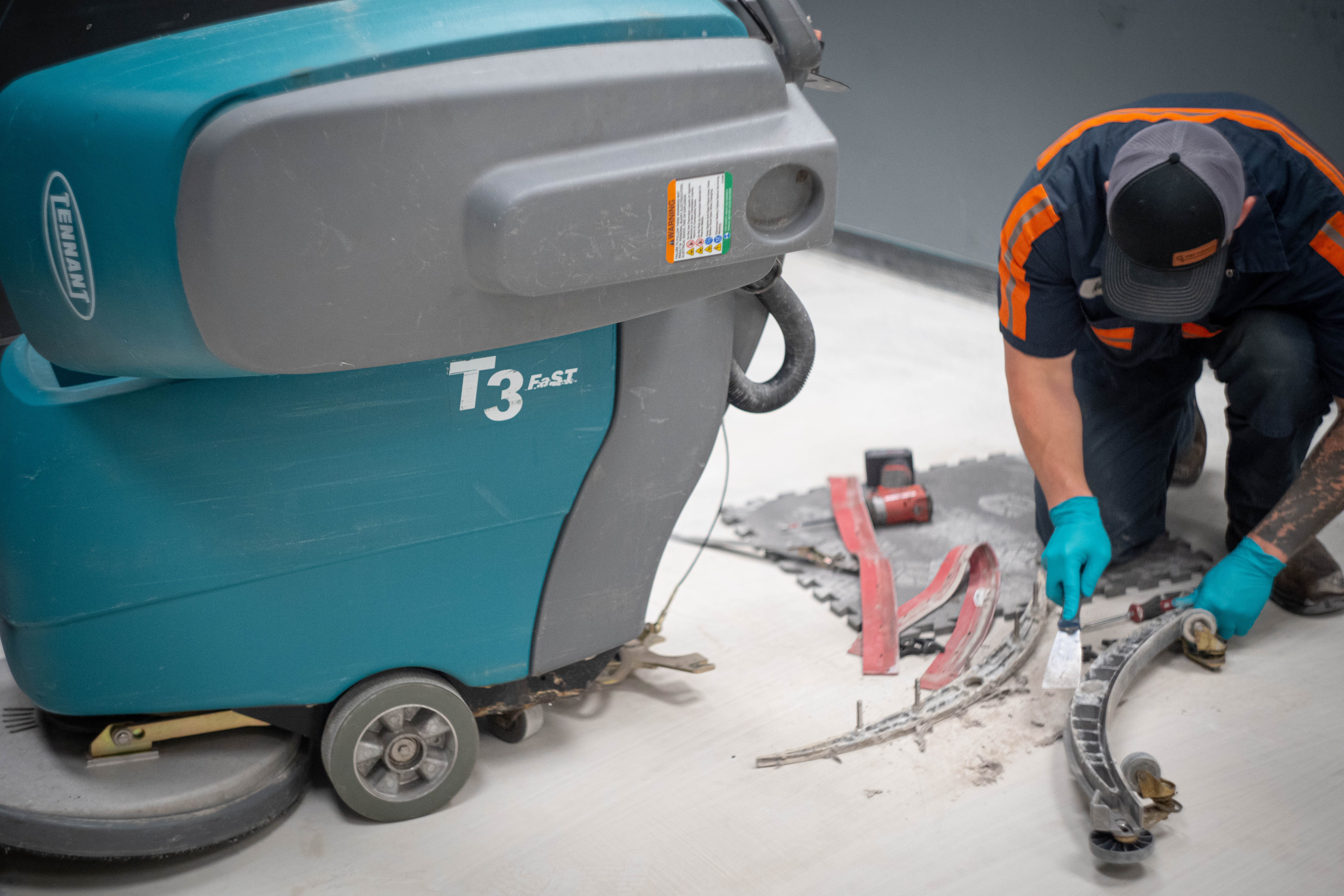 A professional WaySolutions technician meticulously cleans the detached aluminum squeegee housing of a teal Tennant T3 FAST walk-behind floor scrubber. By removing built-up debris and grime before installing high-quality OEM replacement blades, our experts ensure peak water recovery performance, keeping Layton supermarket floors dry and safe for heavy foot traffic.