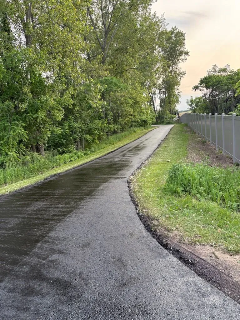 Long narrow asphalt driveway bordered by trees and fencing, showcasing smooth pavement and professional installation quality.