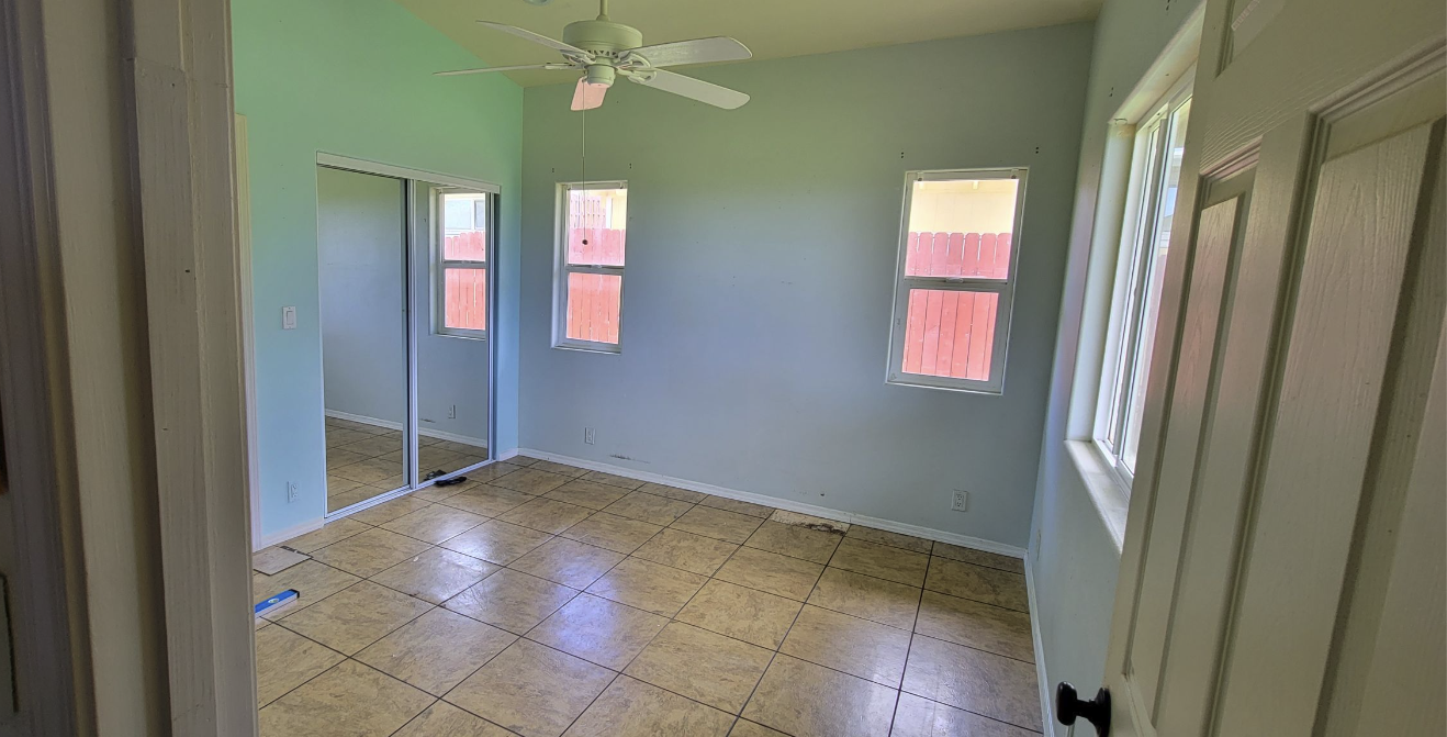 A small, empty bedroom with a vaulted ceiling, painted light green/blue, and yellowish-brown tiled flooring. It features a white ceiling fan, two windows, and a mirrored closet door.