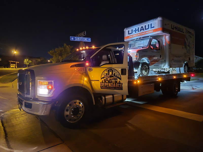 A tow truck from Dependable Towing & Recovery Services transports a U-Haul truck at night, with flashing lights illuminating a residential street, showcasing reliable roadside assistance and vehicle transport services.