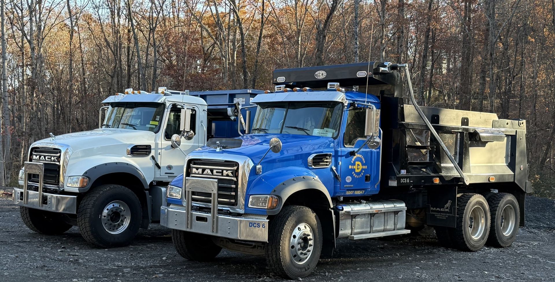Two large Mack dump trucks, one white and one blue with Dinsmore Construction Services branding, are parked side by side on a gravel lot surrounded by tall trees.