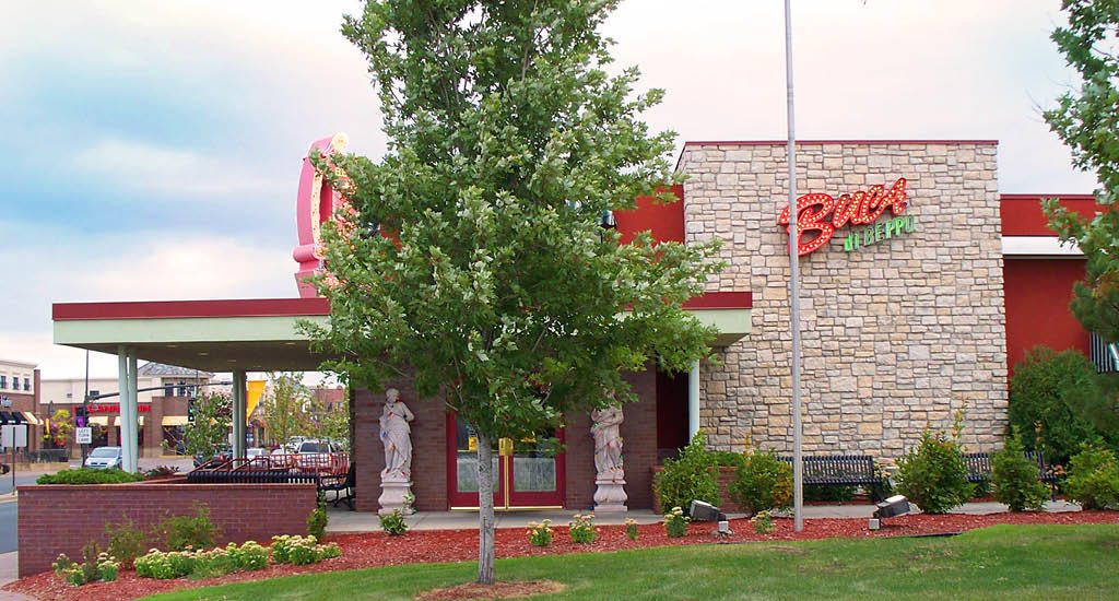 Buca di Beppo Maple Grove side view with greenery, sidewalk, benches, and Buca sign.