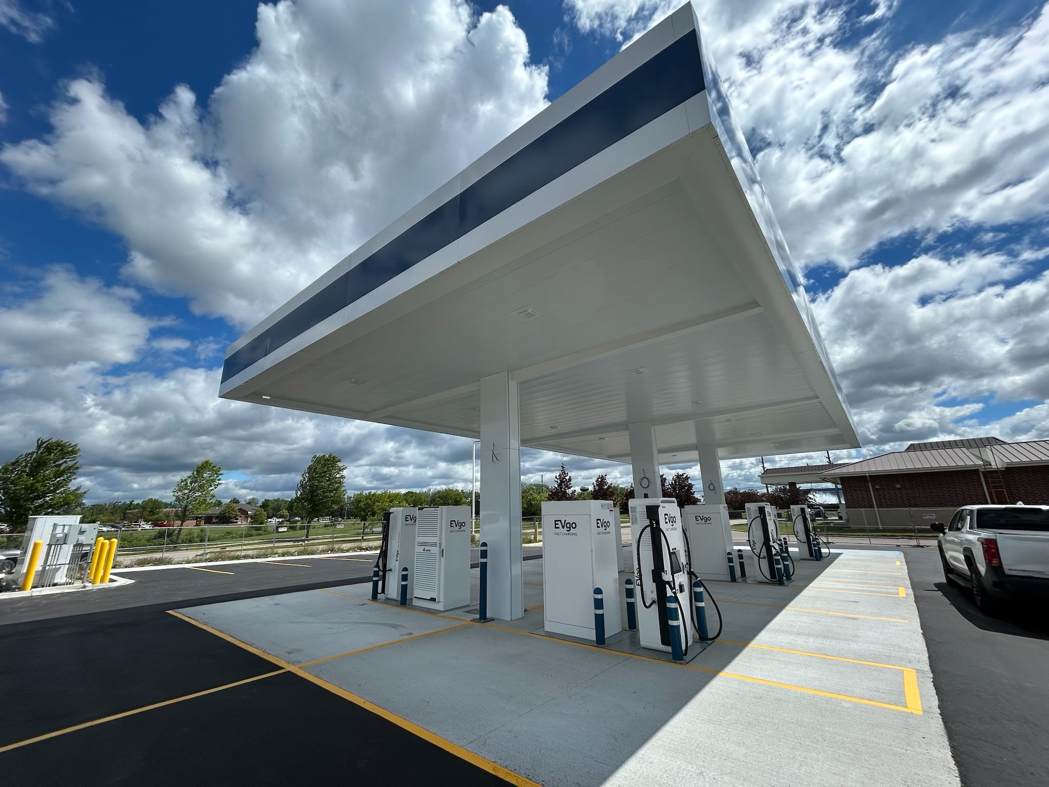 EVgo fast charging station with multiple fast chargers arranged in rows beneath a large white canopy with navy blue trim, set against a bright blue sky with scattered clouds.