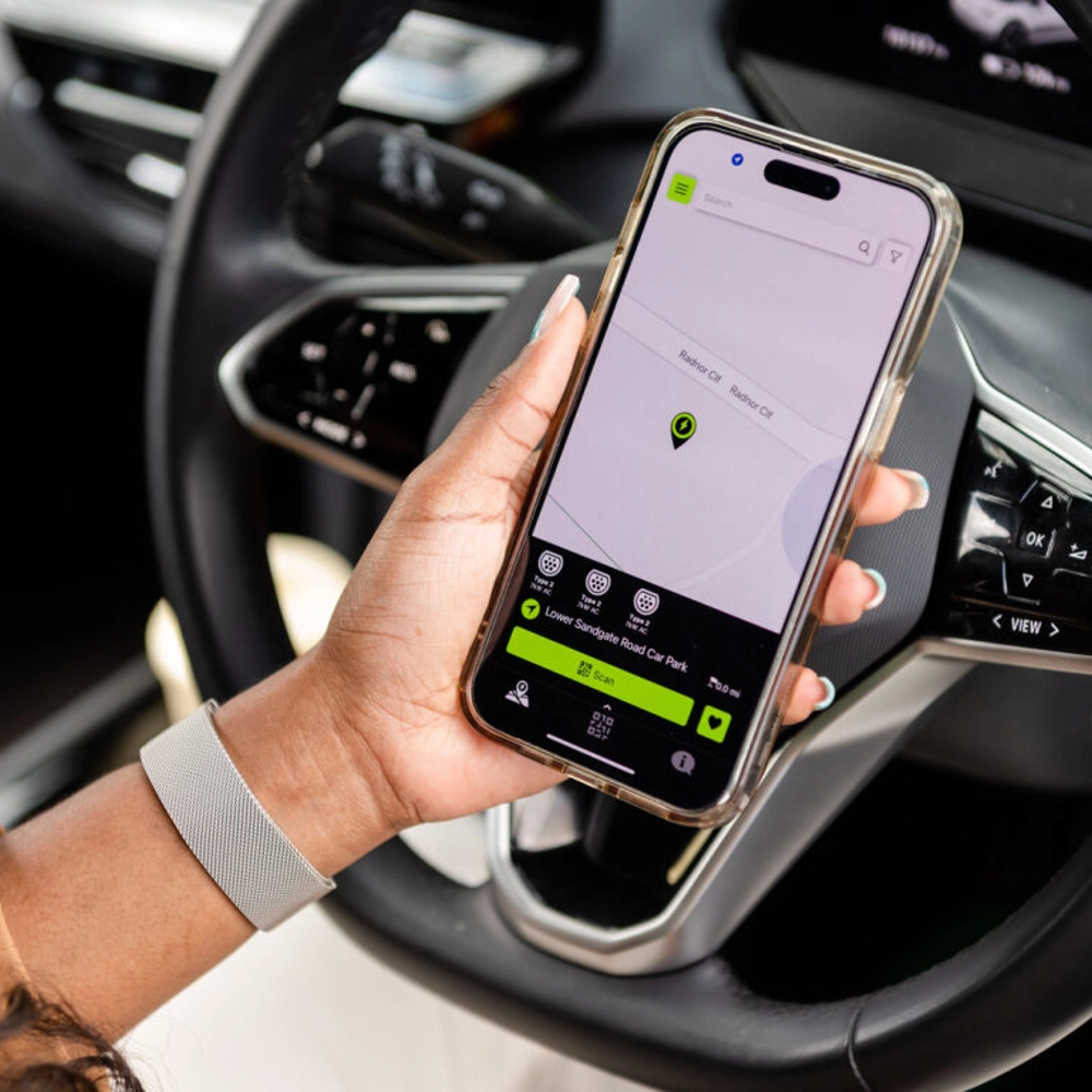 A person sitting in a car holds a smartphone displaying a navigation or parking app on the screen, showing a map with a location pin. The steering wheel and dashboard are visible in the background.