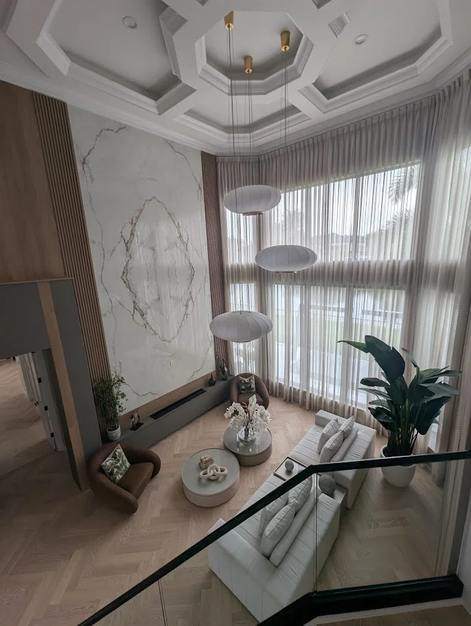 A high-angle shot of a luxurious, modern living room with soaring ceilings and large windows. The room features a statement marble wall, a long white sectional sofa, two brown armchairs, and a modern fireplace. Several large, oval-shaped pendant lights hang from the ceiling, and a tall green plant adds a touch of nature. The flooring is a light-colored herringbone wood.