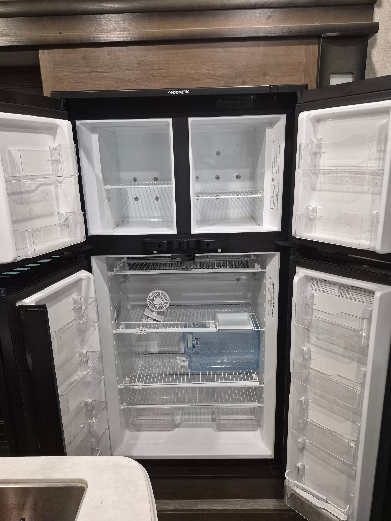 Open Dometic RV refrigerator and freezer with multiple doors, white interior shelves, transparent bins, small fan, ice tray, and water container, showing clean storage compartments during appliance inspection or service.