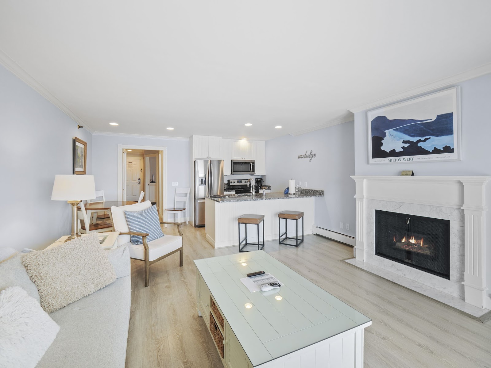 A bright and airy living room and kitchen area with a fireplace, a sofa, a coffee table, and a kitchen island with stools. A large piece of abstract art hangs above the fireplace. The room is decorated in neutral tones with accents of blue and white.