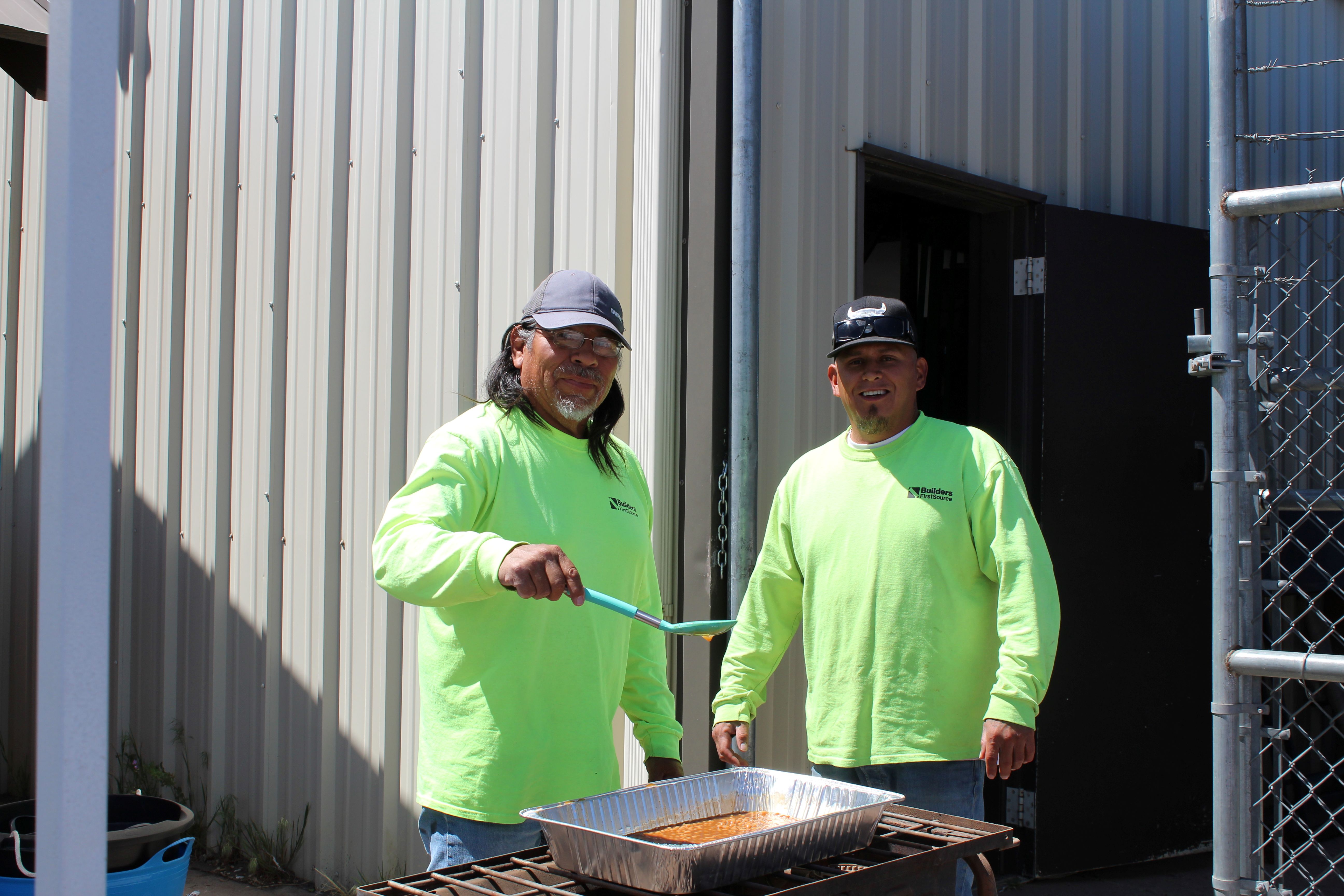 Two people wearing bright green long-sleeve shirts stand at an outdoor grill beside a metal building. One person uses a ladle to stir food in a large foil pan set on the grill, while the other stands beside them near an open door and chain-link gate.