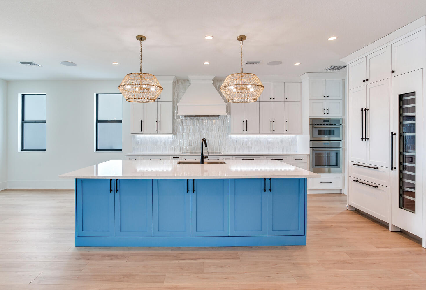 Bright coastal-style kitchen with a blue island, white cabinetry, pendant lighting, wood flooring, and an open-concept luxury layout.