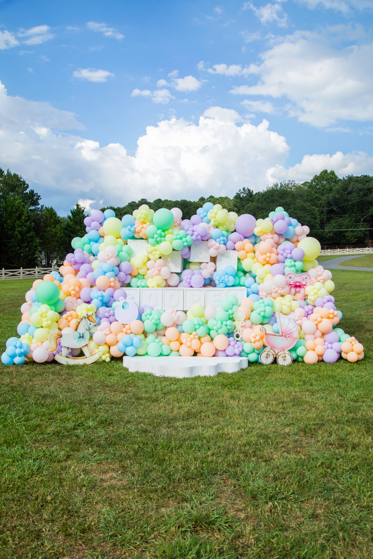 Whimsical outdoor baby shower backdrop featuring pastel balloon wall, soft rainbow colors, white pedestal, and playful baby-themed props, set on grassy field under blue sky for cheerful celebration photos.