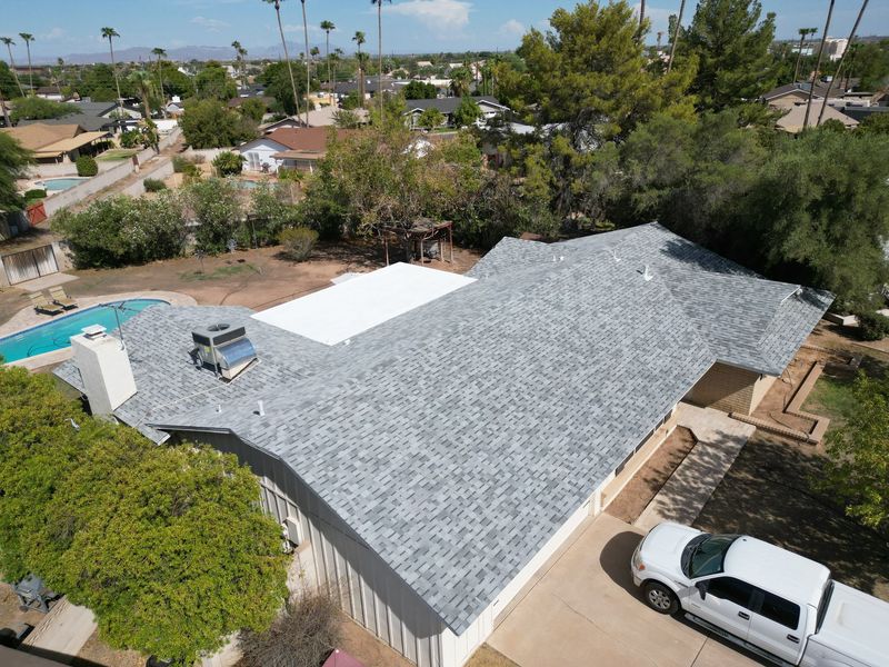 A high-angle drone shot captures a sprawling residential property featuring a freshly installed light gray shingle roof. The house includes a white flat-roof section, a backyard pool, and tall trees.