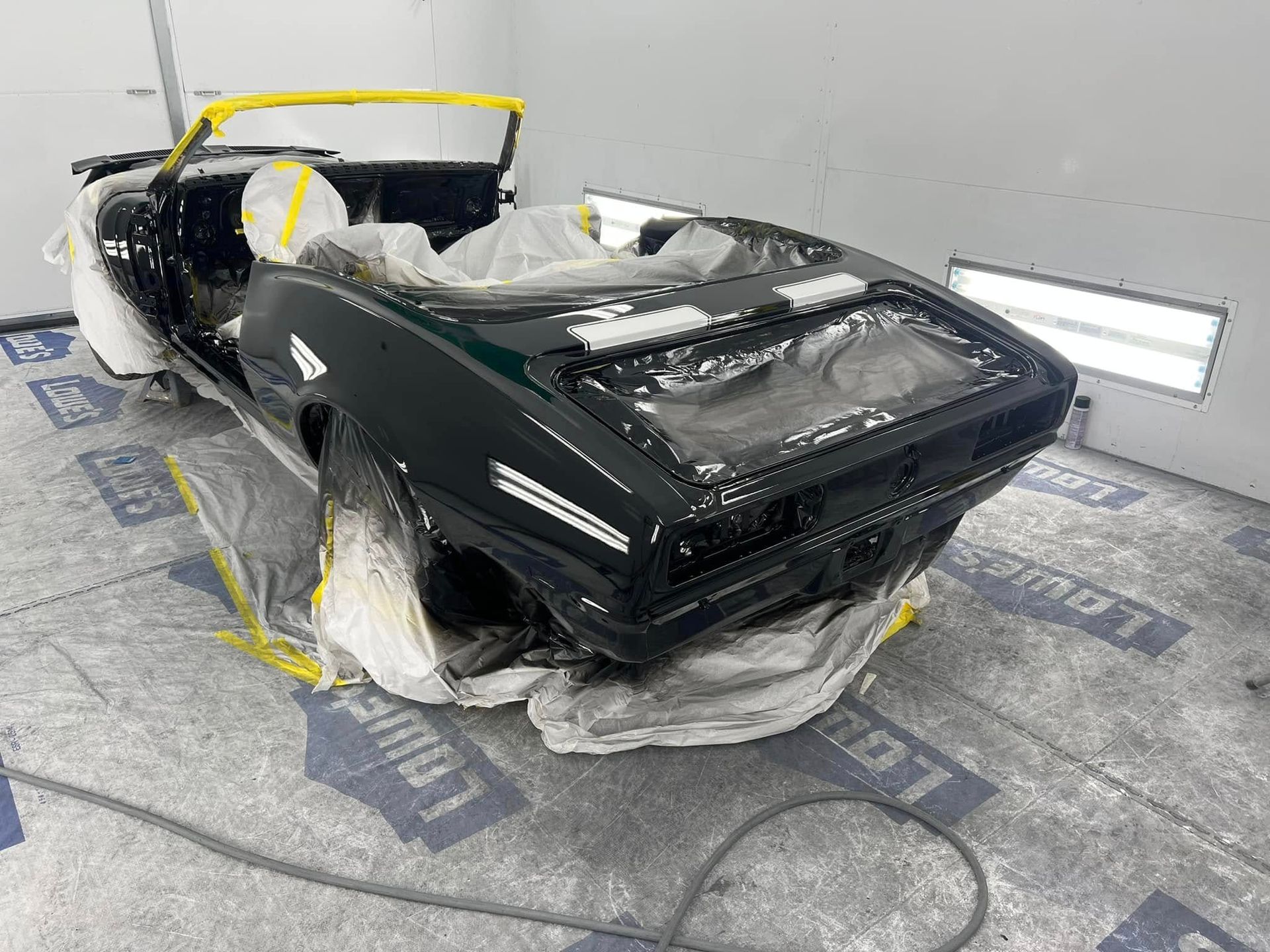 A restored Camaro convertible features a freshly applied black finish and white accent stripes. The vehicle remains masked while the paint cures inside a controlled booth.