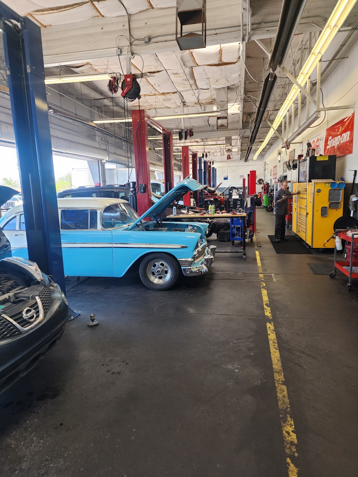 A mechanic shop bay with several cars being worked on, including a vintage blue and white car with its hood open and a newer dark-colored car with its hood open. The bay is equipped with car lifts, tool chests, and various automotive equipment. A mechanic in a gray shirt and dark pants is visible near a yellow tool chest. A red Snap-on banner hangs on the wall.