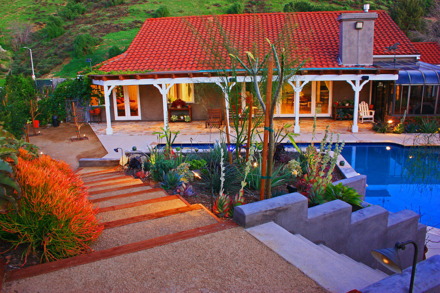 Tuscan-style villa with red tile roof, lush hillside garden, stone paths, desert plants, and a blue pool, creating a warm, tranquil retreat.
