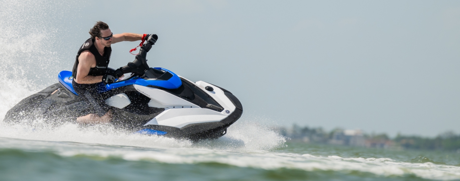 A man is riding a jet ski through the water, creating a splash as he turns. He is wearing sunglasses and a life vest, and the jet ski is blue and white.