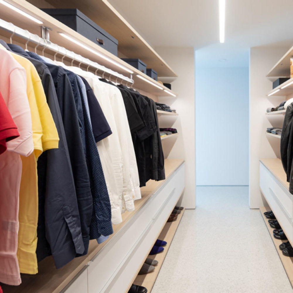 Modern walk-in closet with neatly organized clothes hanging on both sides, shelves holding storage boxes, and shoes lined up on the lower shelves. Bright overhead lighting illuminates the clean, minimalist space.