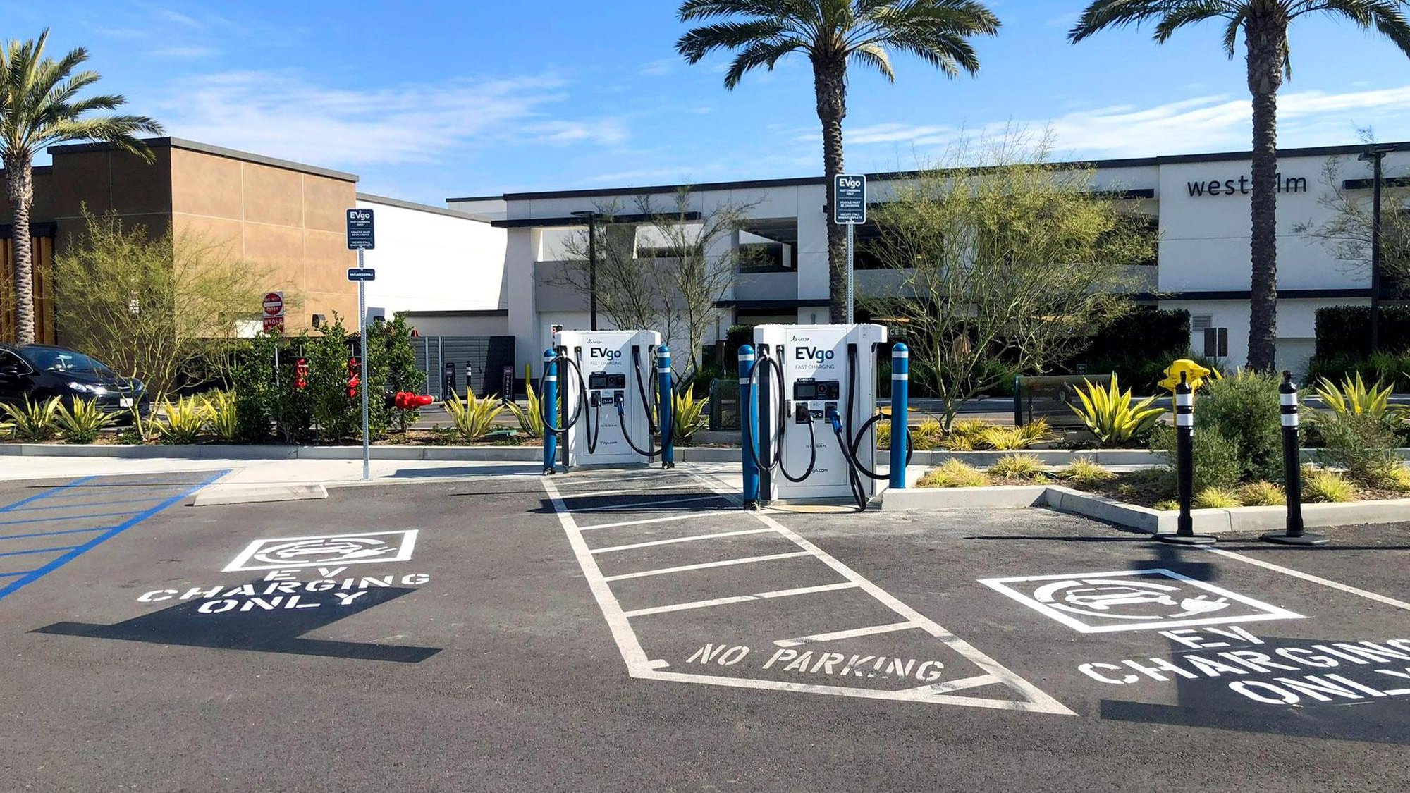 Several EVgo fast charging units installed in marked parking spaces at a public charging station, with EVgo logos on the chargers, protective bollards around each unit.