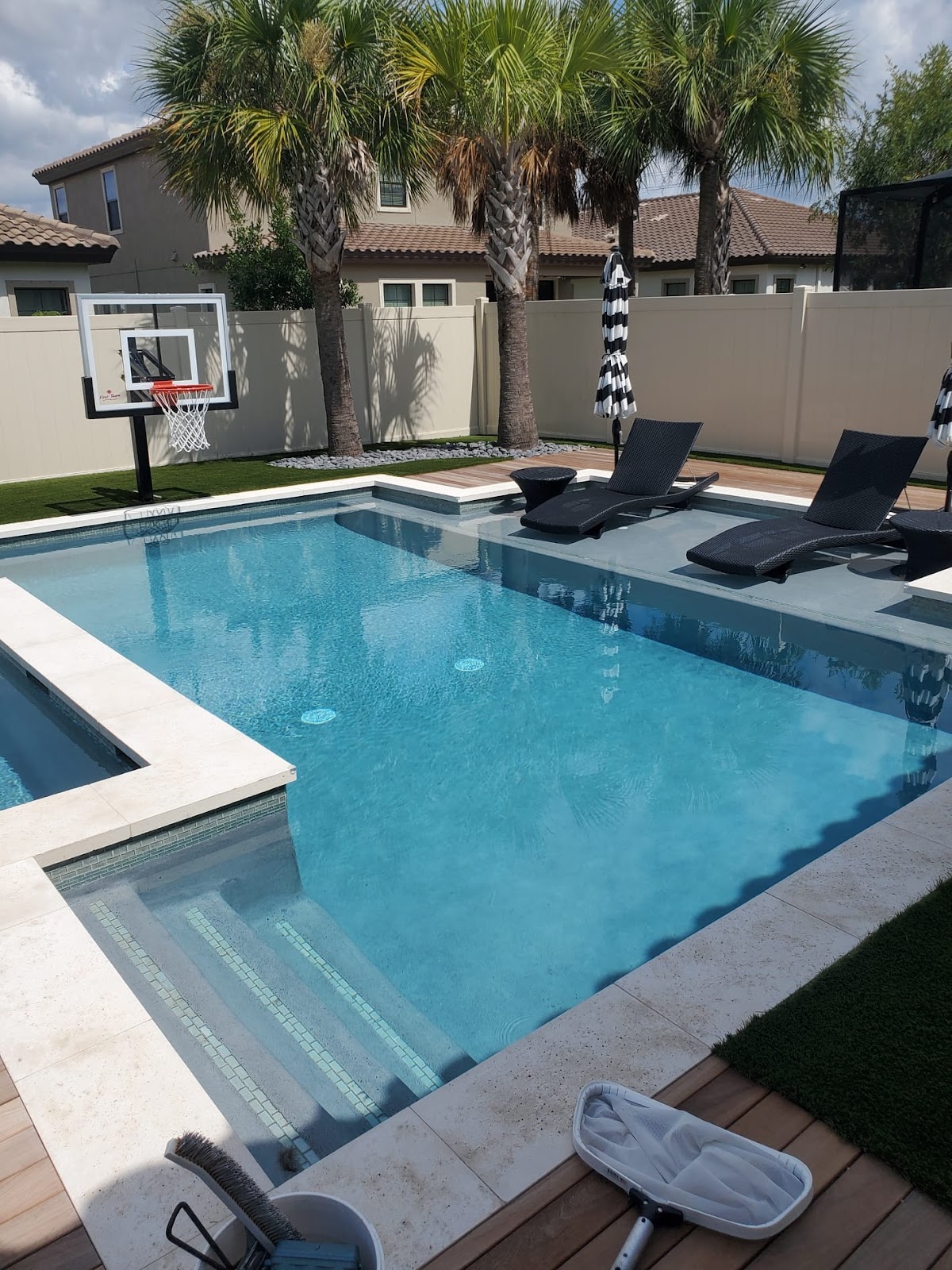 Backyard swimming pool with a shallow entry area, lounge chairs, palm trees, and a basketball hoop beside the pool.