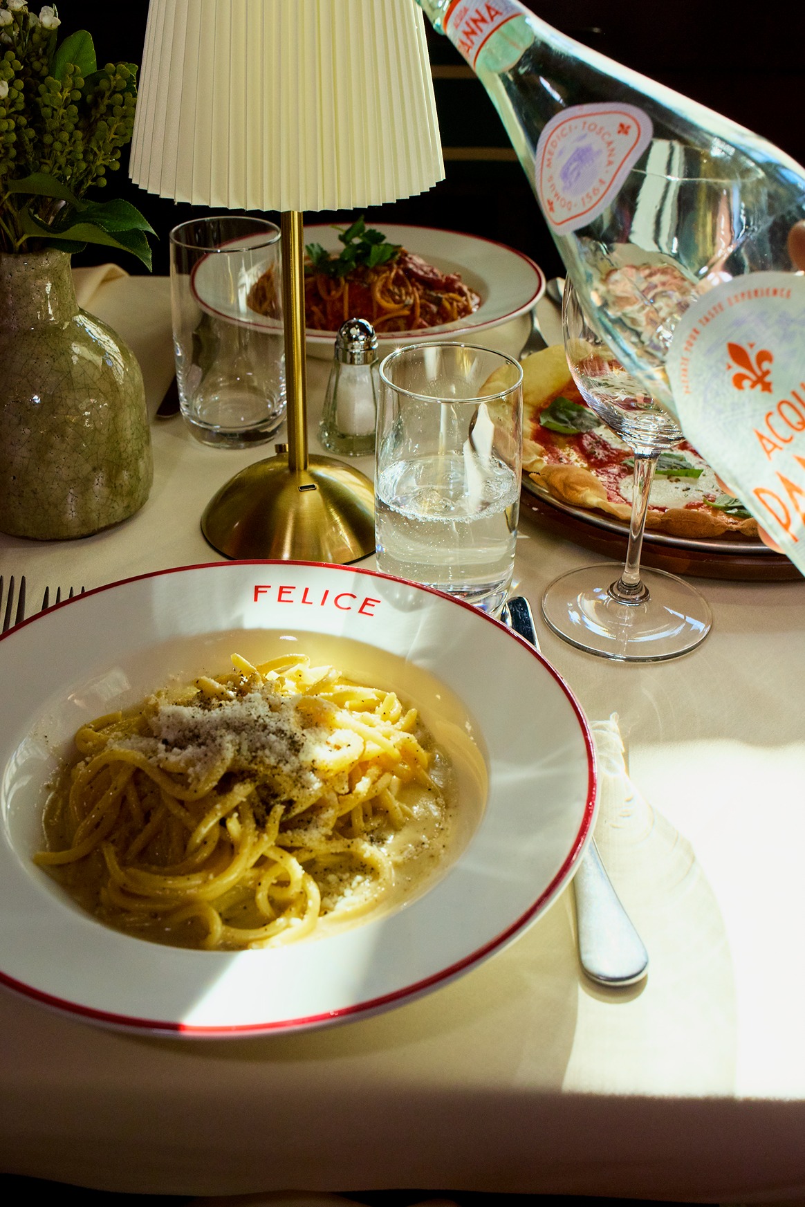 Creamy spaghetti topped with cracked black pepper and pecorino sits in a white bowl. A bottle of Acqua Panna pours into a glass nearby under warm lamp light.