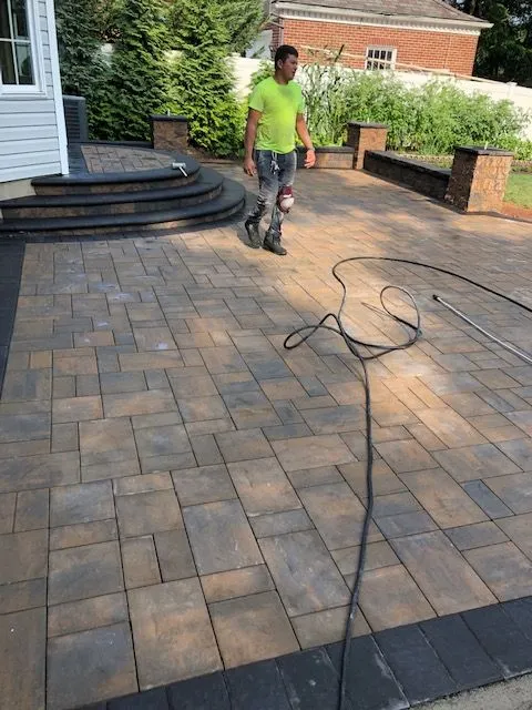 A man in a bright green t-shirt and jeans walks across a newly paved stone patio with a black border. He is wearing knee pads and has a hose lying on the patio in front of him.
