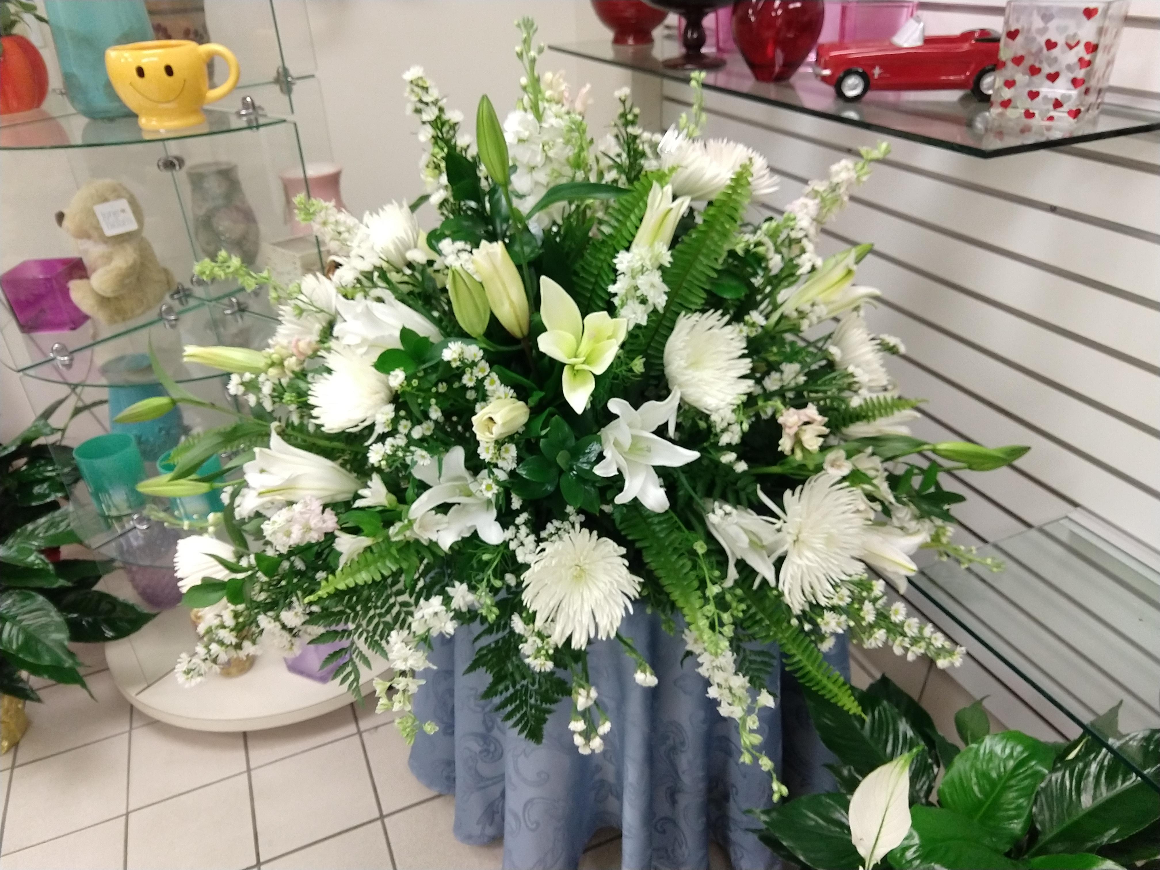 A casket cover for funeral services designed in a half circle that sits on the casket. Flowers include white Asiatic lilies, white stock, white larkspur, white gerbera daisies, white mums, and lush foliage.