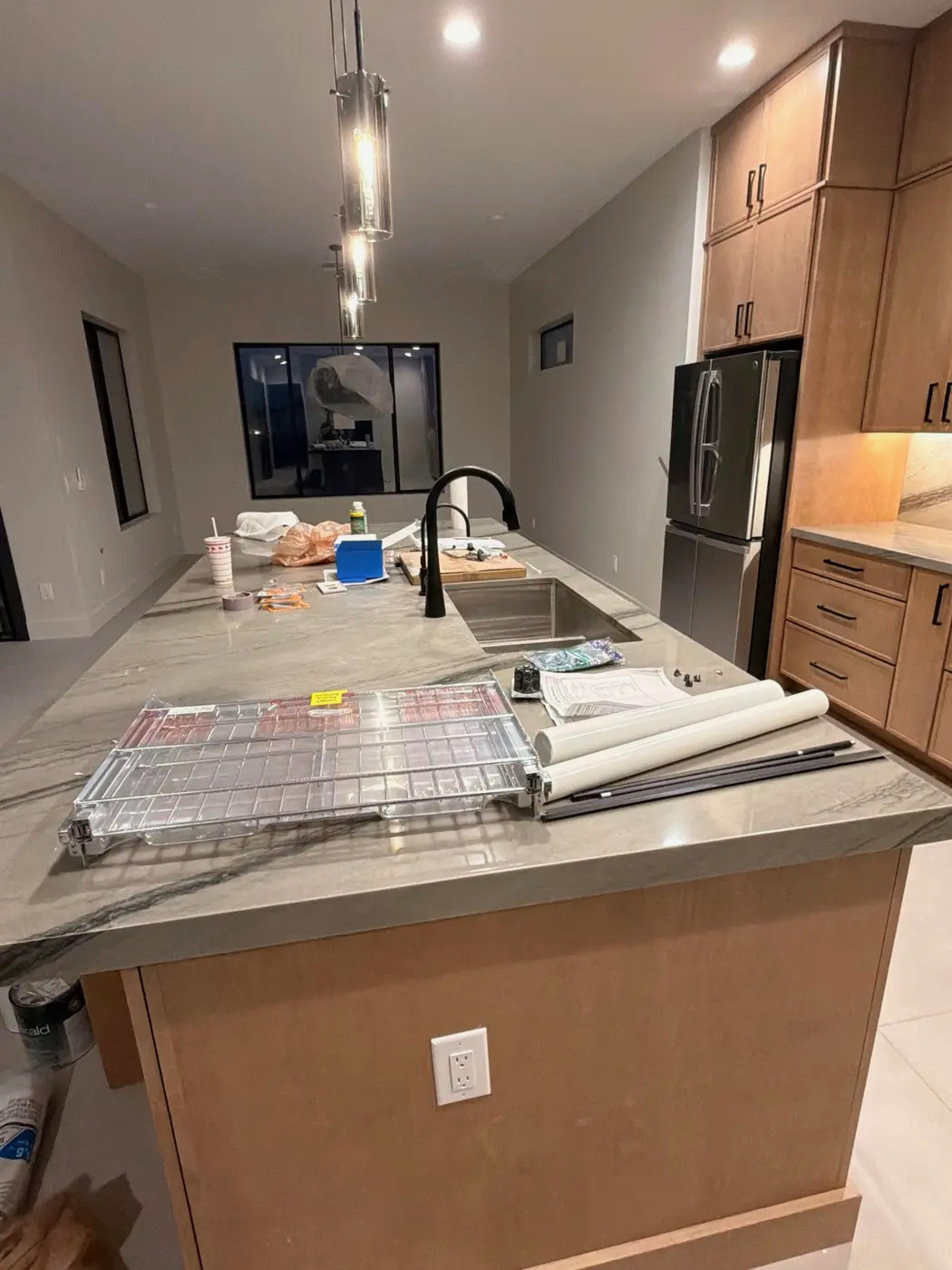 A modern kitchen island is shown with a countertop made of gray and white marble. Several items are placed on the countertop, including a metal rack, rolled-up papers, and a black faucet over a sink. To the right, a stainless steel refrigerator and wooden cabinets are visible. The kitchen appears to be under renovation or recently cleaned after construction, with some construction materials like plastic wrap and a can of paint visible on the floor.