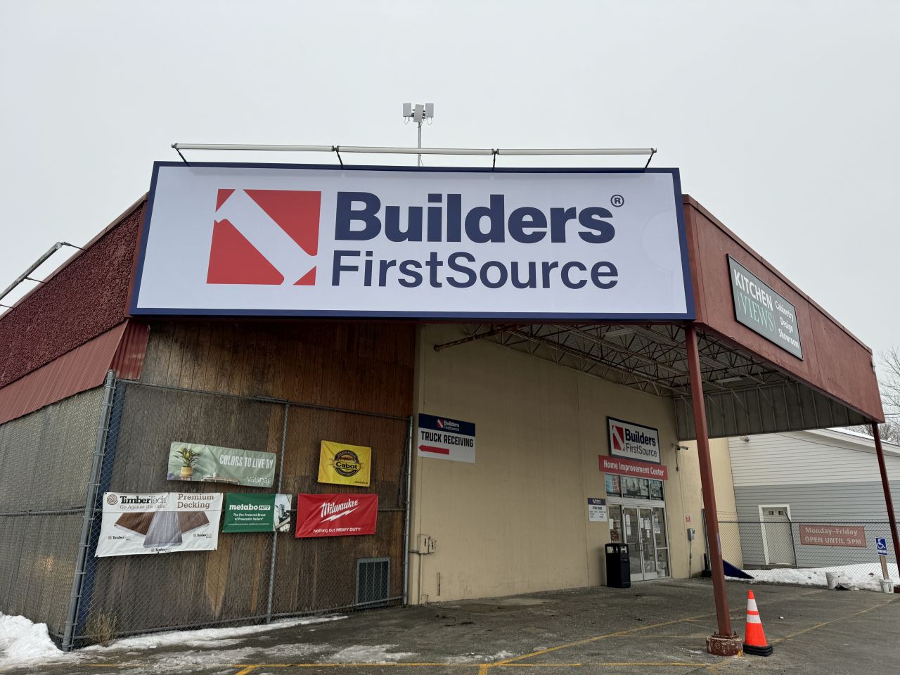 Exterior of a Builders FirstSource building materials location with large branded signage, featuring a covered front entrance, parking area, delivery access, and winter conditions outside a professional lumber and building supply facility.