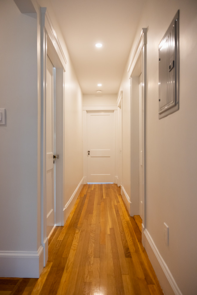 A straight-on, eye-level, medium shot of a well-lit, narrow hallway with white walls, white trim, and a polished hardwood floor. Several closed white doors are visible along the hallway, some with ornate doorknobs. A metal electrical panel is mounted on the right wall. The hallway recedes into the distance, with a bright light at the end, suggesting an exit or another room. The overall impression is one of clean, modern, and well-maintained interior space.