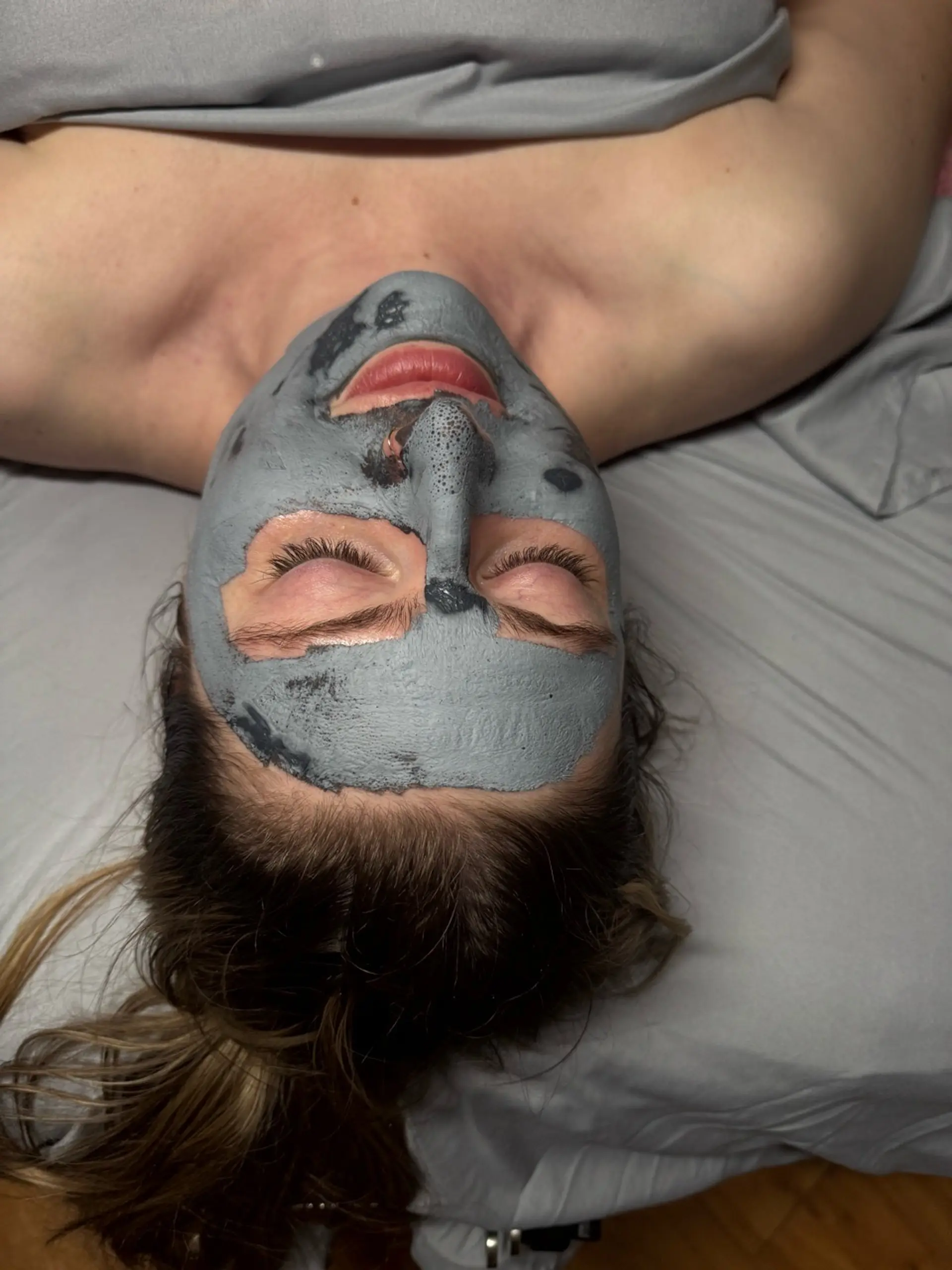 A person's face, tilted upwards, is covered in a gray charcoal mask, with their eyes closed and lips slightly parted. The mask covers their entire face, leaving only the lips and eyelids exposed. The person is lying on a gray surface, possibly a bed or massage table, and wearing a gray top. Their hair is dark brown and pulled back.