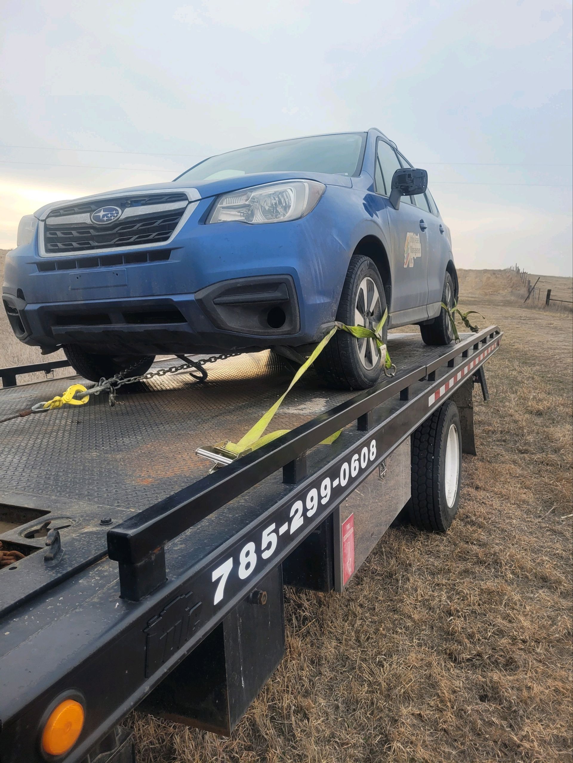 A low-angle photograph shows a blue Subaru SUV mounted on a flatbed tow truck. Bright yellow straps secure the front tires while a phone number is printed on the truck rail.