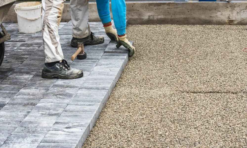 Two workers are laying down gray paving stones next to a bed of gravel. One worker is placing a stone into position with gloved hands, while the other worker, wearing work boots and light-colored pants, stands nearby. A sledgehammer rests on the ground next to the finished paving stones. A white bucket and part of a wheelbarrow are visible in the background. The scene appears to be an outdoor construction or landscaping project.