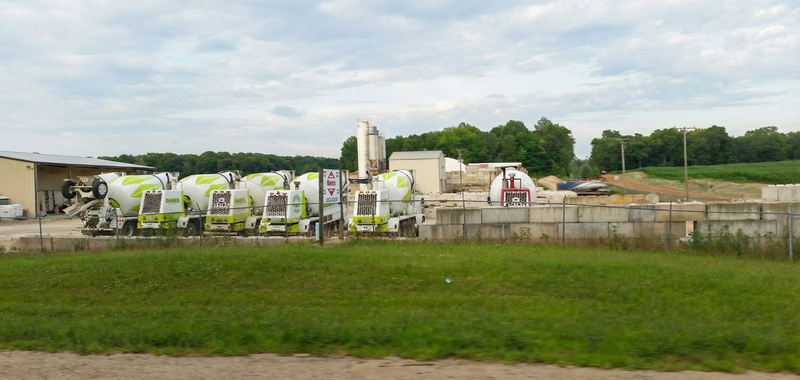 A fleet of lime-green and white Dave's Concrete mixing trucks is parked in a row at an industrial facility, featuring a tall cement silo, storage buildings, and concrete blocks.