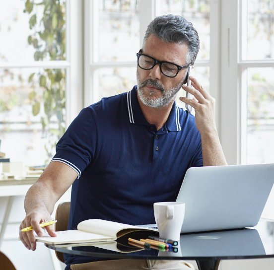 A man with gray hair and a beard, wearing glasses and a dark blue polo shirt, is sitting at a table, talking on his cell phone. In front of him is an open laptop, a notebook, a white mug, and several colored pens. The background shows a window with greenery outside.