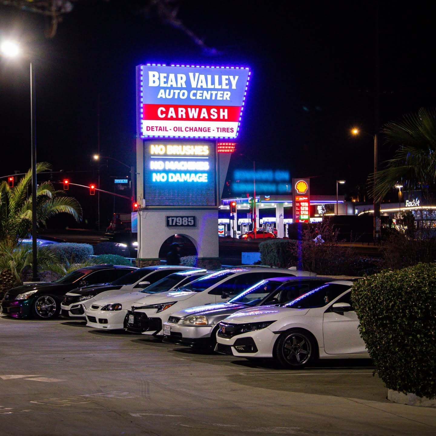 Lineup of modified cars displayed beneath a brightly illuminated automotive center sign during a nig 